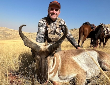 A hunter smiling alongside a trophy pronghorn antelope, with horses saddled in the background on a Wyoming plains hunt.